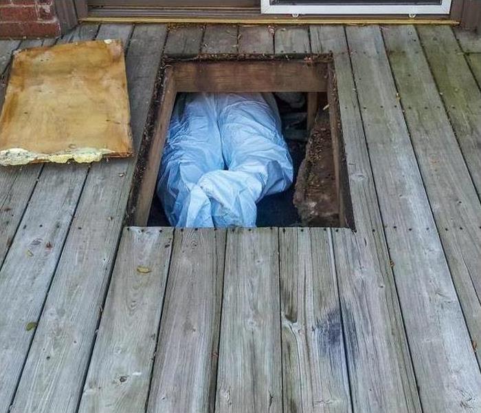 Workman with protective suit crawling under house from crawlspace underneath a wooden deck.